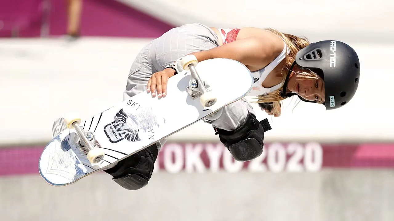 Atleta di skateboard con casco nero durante una manovra aerea in una competizione olimpica, con scritta Tokyo 2020 sullo sfondo