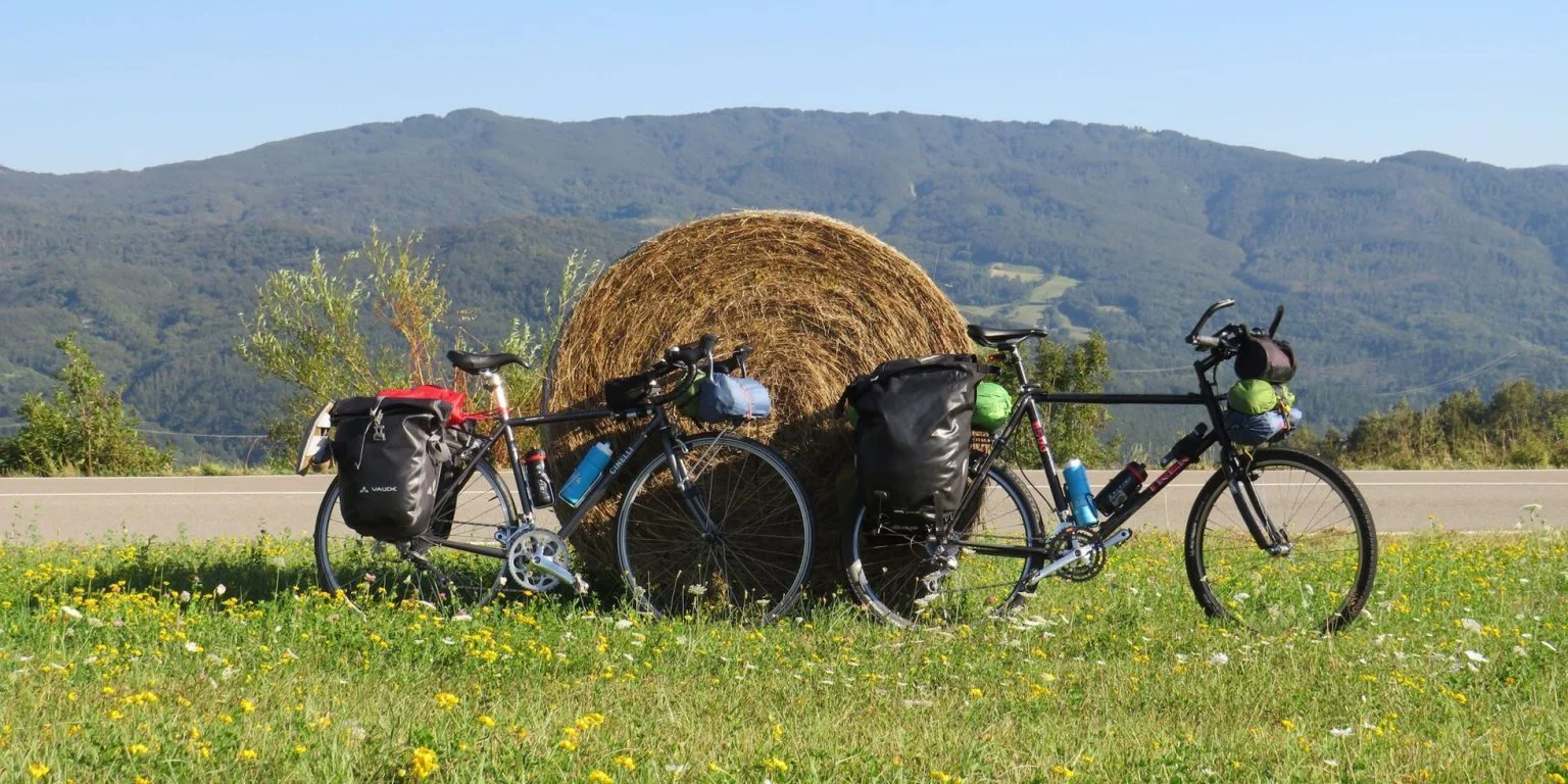 Cicloturista con maglia gialla e casco, in sella a una bicicletta con borse da viaggio, lungo una strada di campagna tra i campi