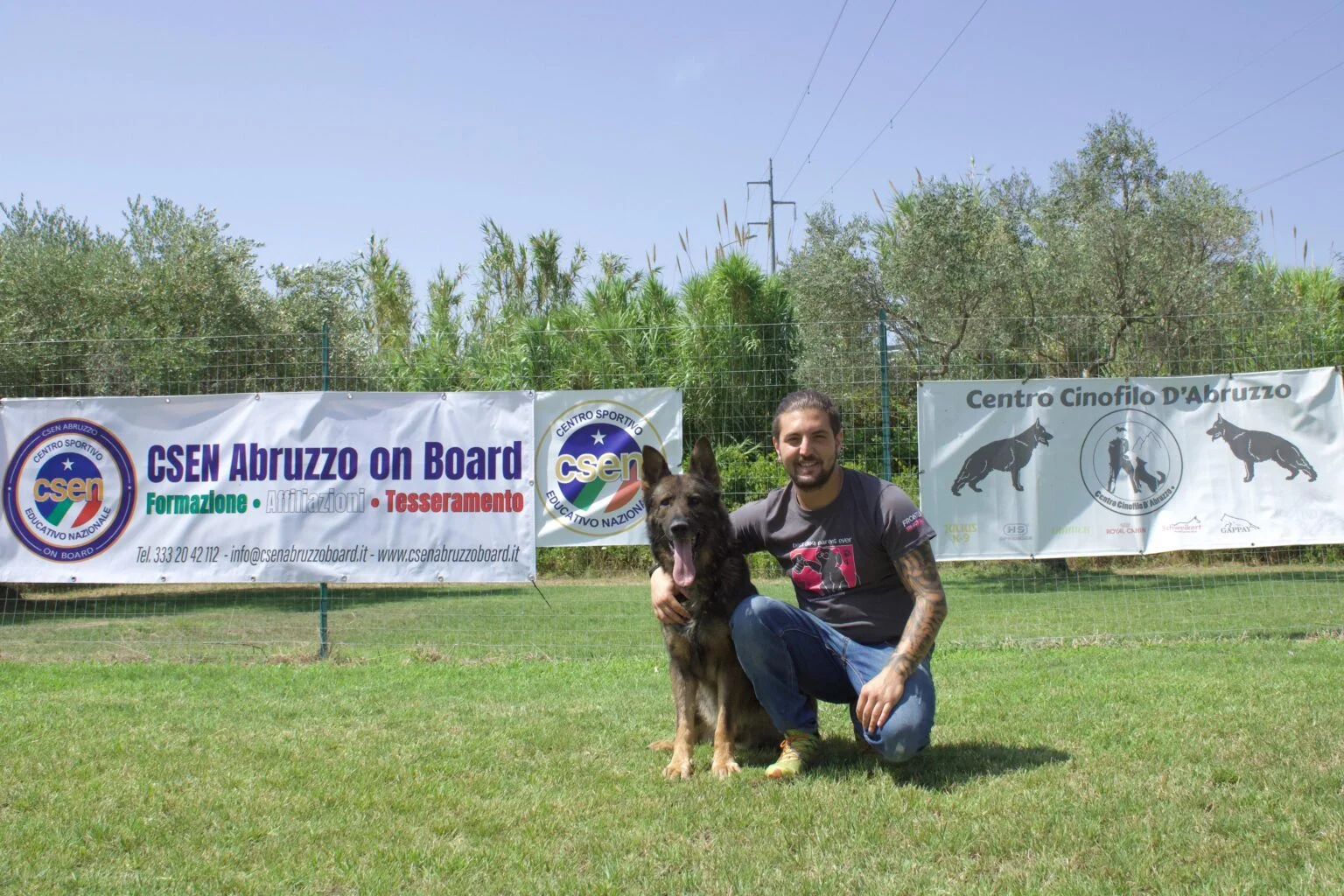 Uomo accanto a un pastore tedesco seduto sul prato, davanti agli striscioni CSEN Abruzzo On Board e Centro Cinofilo d'Abruzzo