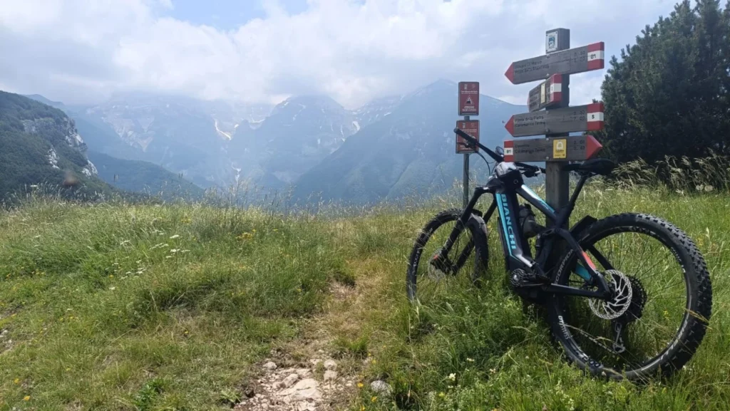 Mountain bike elettrica Bianchi accanto alla segnaletica escursionistica, con montagne e prati sullo sfondo