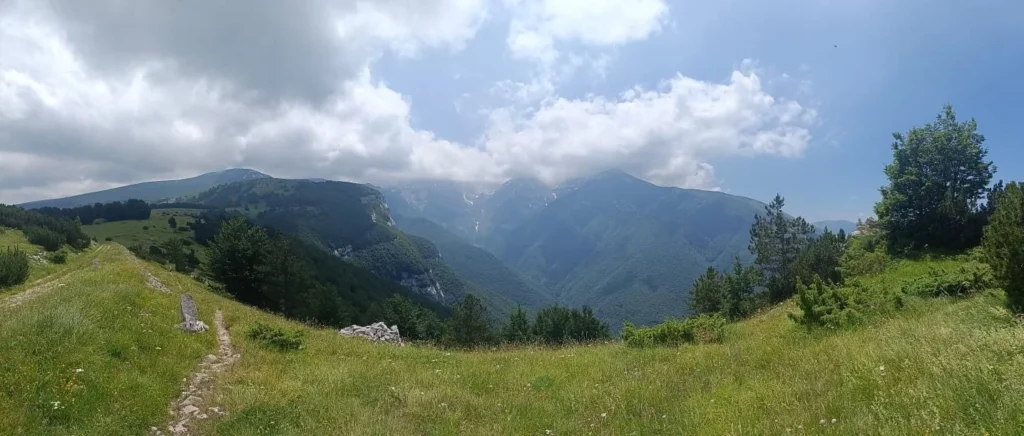 Sentiero tra i prati di montagna, con boschi e rilievi parzialmente coperti dalle nuvole