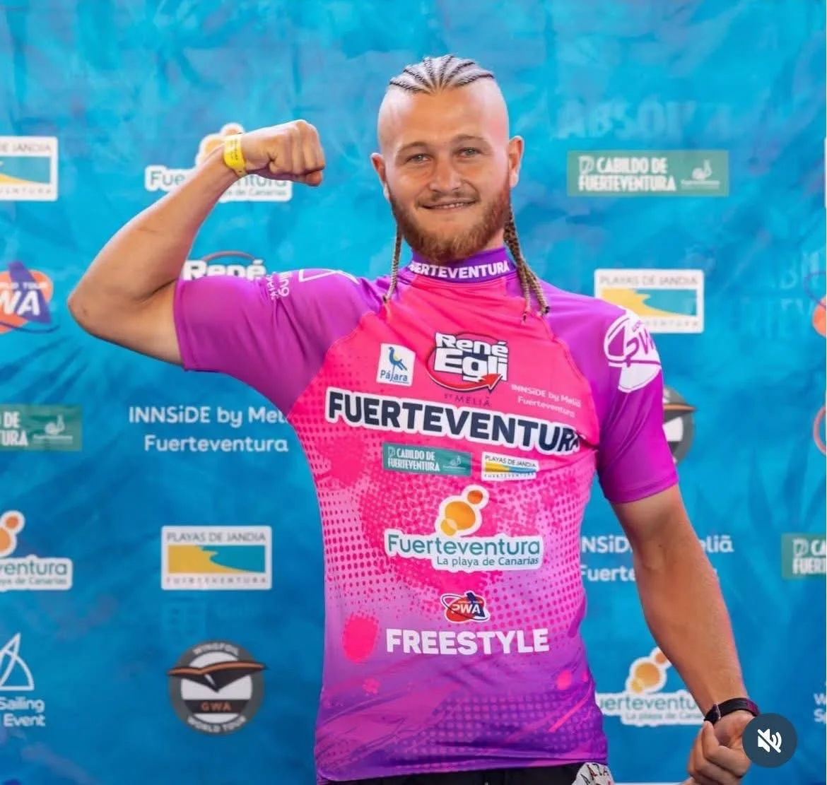 Nicolò Di Marco con maglia rosa da competizione solleva un braccio davanti al pannello degli sponsor di Fuerteventura