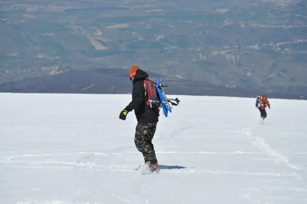 Snowboarder in discesa sulla neve con ciaspole fissate allo zaino e un altro praticante più a valle
