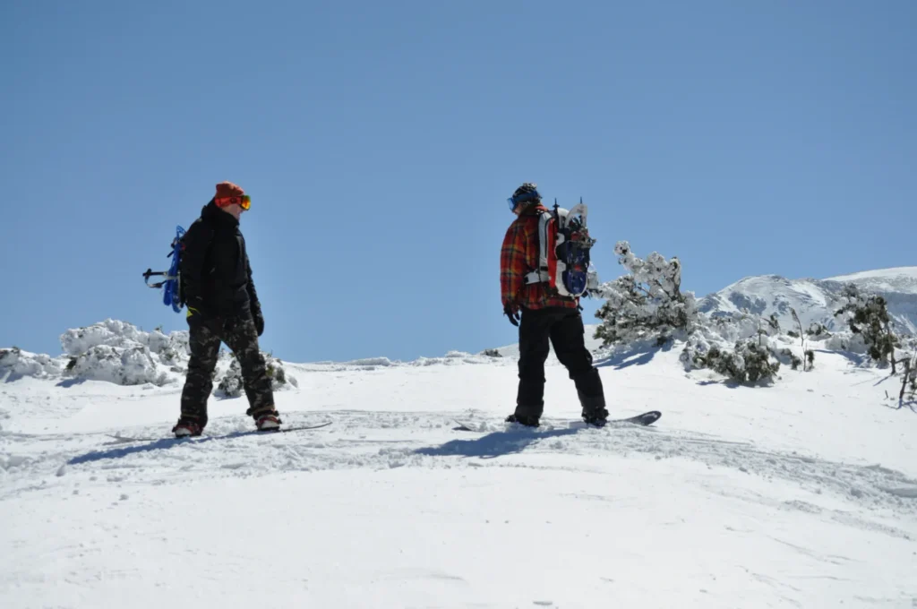 Due snowboarder fermi sulla neve con ciaspole fissate agli zaini, tra alberi innevati e montagne