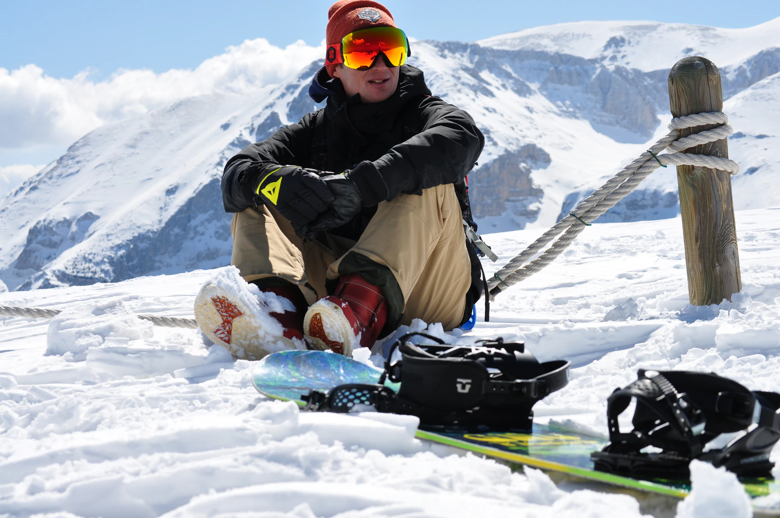 Snowboarder seduto sulla neve con maschera da sci e tavola da snowboard in primo piano, sullo sfondo montagne innevate