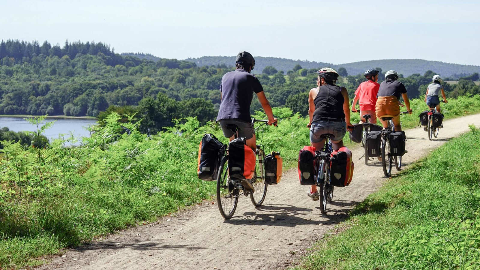 Gruppo di cinque cicloturisti con biciclette e borse da viaggio lungo un percorso sterrato vicino a un lago