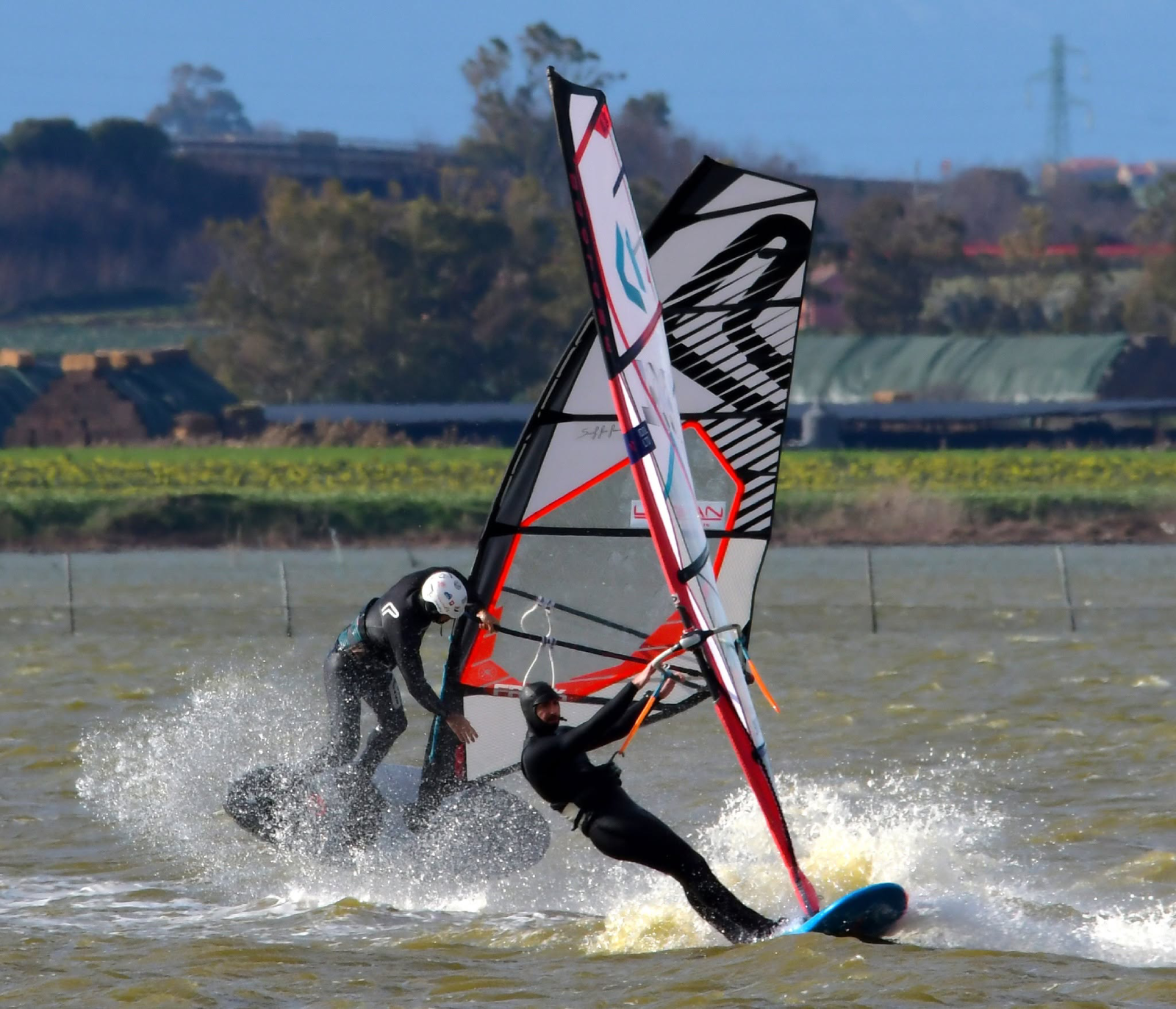 Due windsurfer con mute nere impegnati in manovre ravvicinate sull'acqua, tra vele e spruzzi